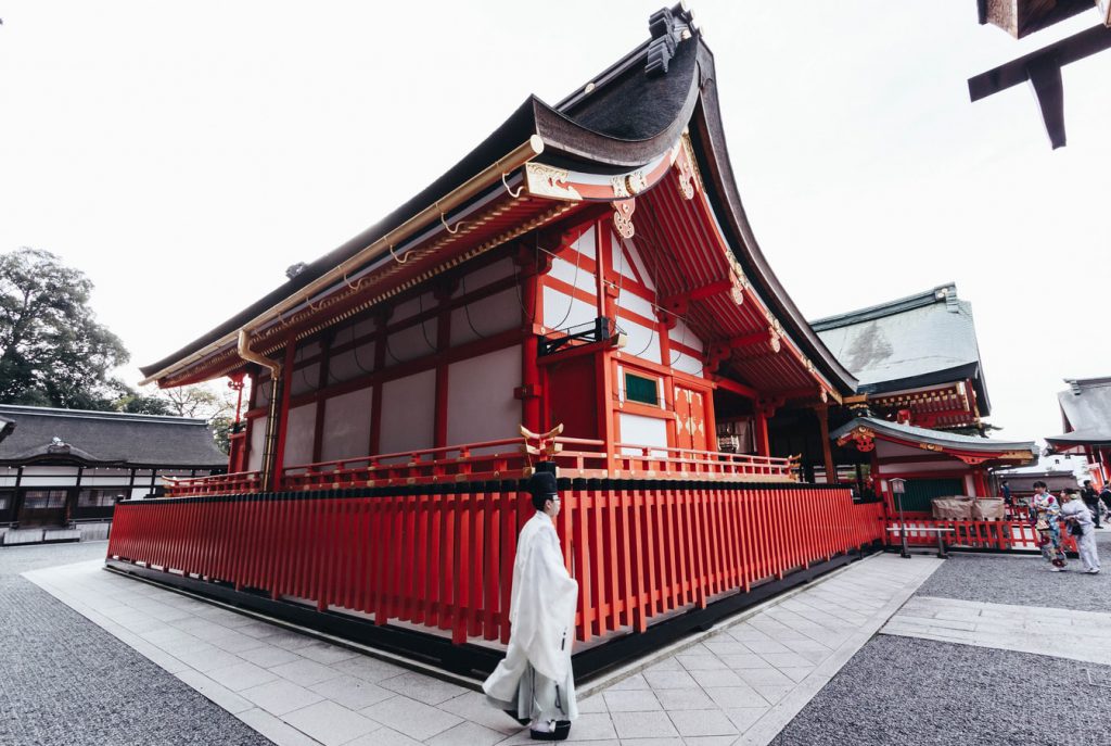 A Japanese priest dressed in white is standing in front of a red shrine