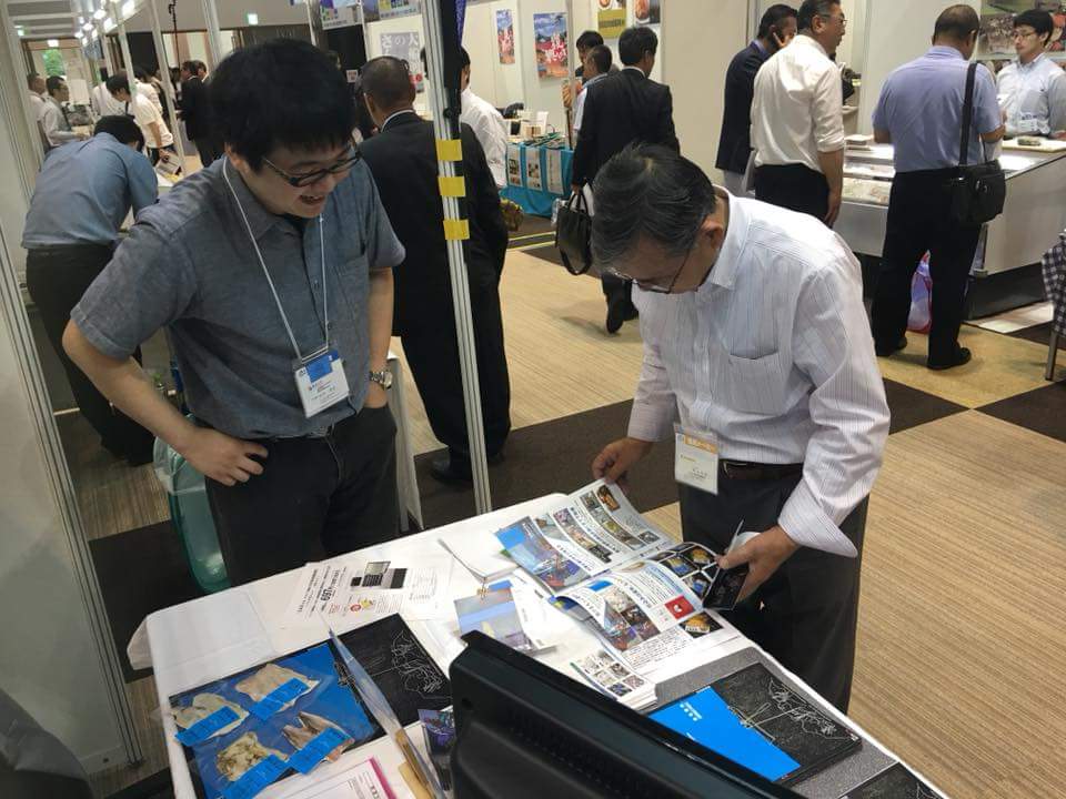 Doctor Suzuki at a booth, talking to someone who is looking at a pamphlet