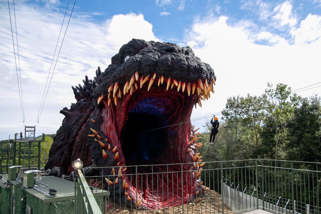 Enormous Godzilla head with its mouth open. Someone on a zip line is about to enter it.