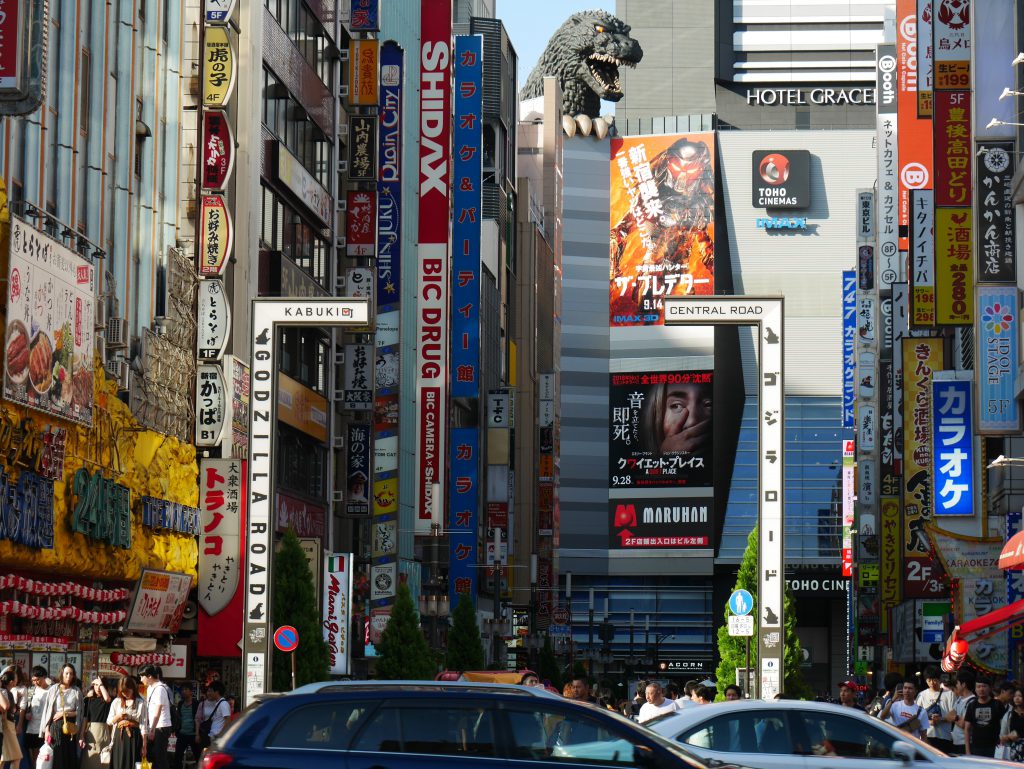 A street with huge buildings and colorful neon lights. At the back, a real-sized Godzilla head and claws on top of Toho Cinemas