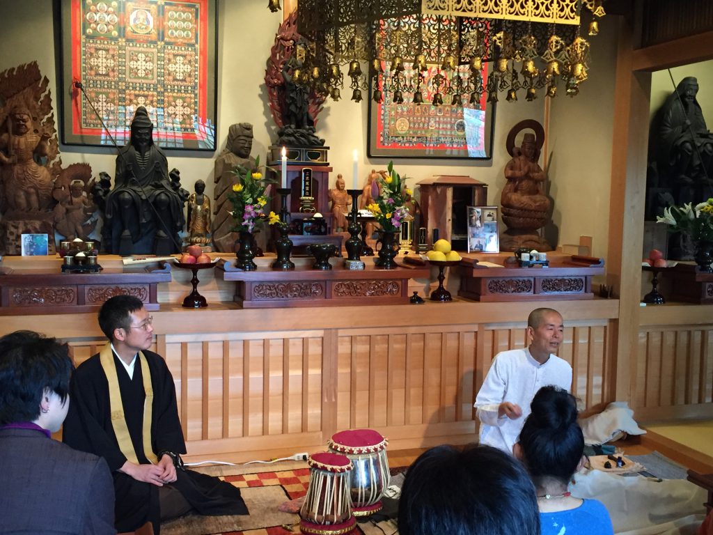 Many people are sitting on the floor inside a temple. Several Buddhist statues can be seen on the altar.