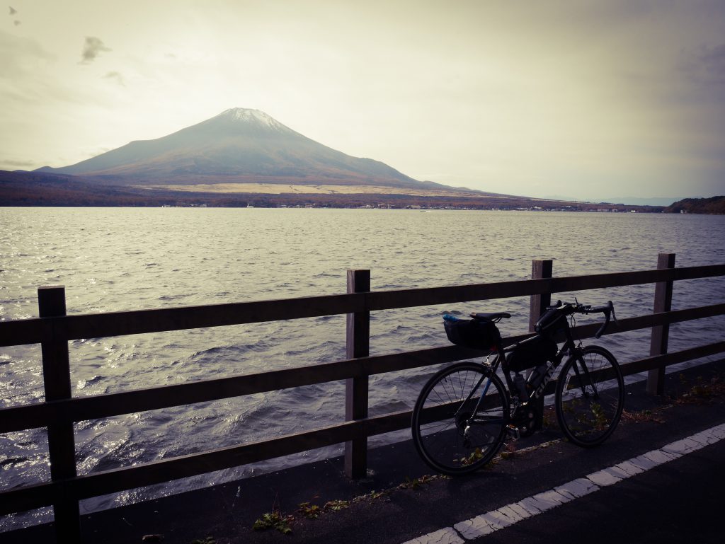 A bicycle rests against a fence along a road. Behind the fence, a lake. On the other side of the shore, far away, is Mount Fuji.