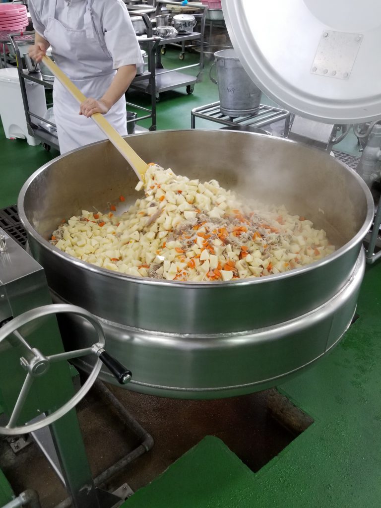 Someone cooking large quantities of vegetables in a pot so huge a person could sit inside