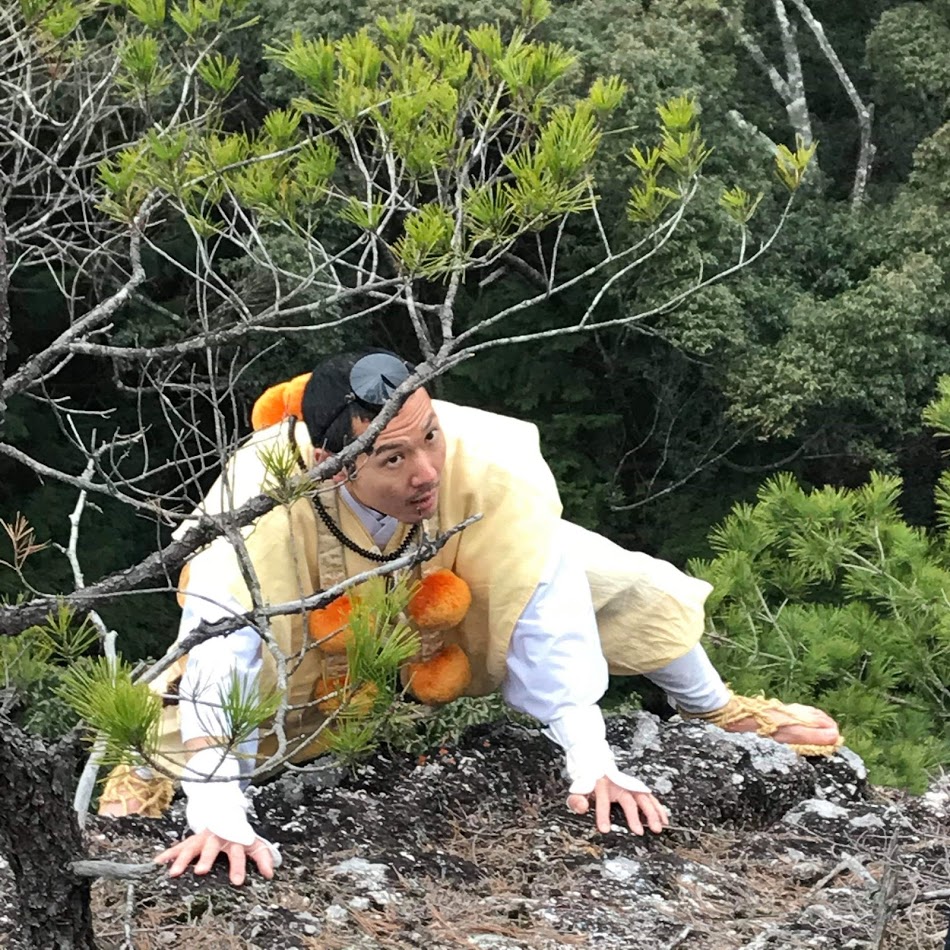 Mr. Tani climbing a rock dressed in the Shugendo priest outfit: white and beige clothes. He has a sort of hat on his forehead, pompons around his neck and straw sandals