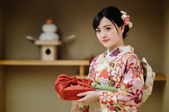 A Japanese woman is dressed in a New Year kimono. A kagami mochi is displayed on a shelf behind her.