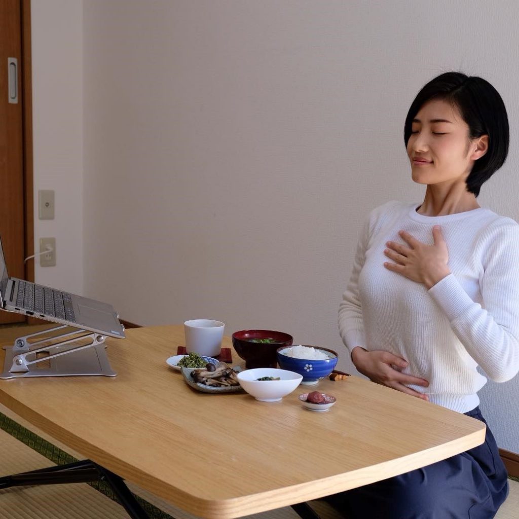 A Japanese woman is sitting in front of  food put on a table. She has her eyes closed and her hand on her heart.
