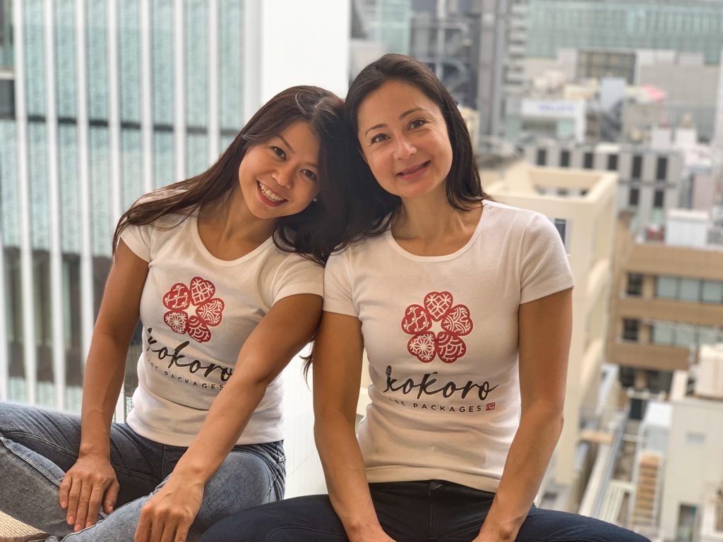 Two women wearing 'Kokoro Care Packages' T-shirts
