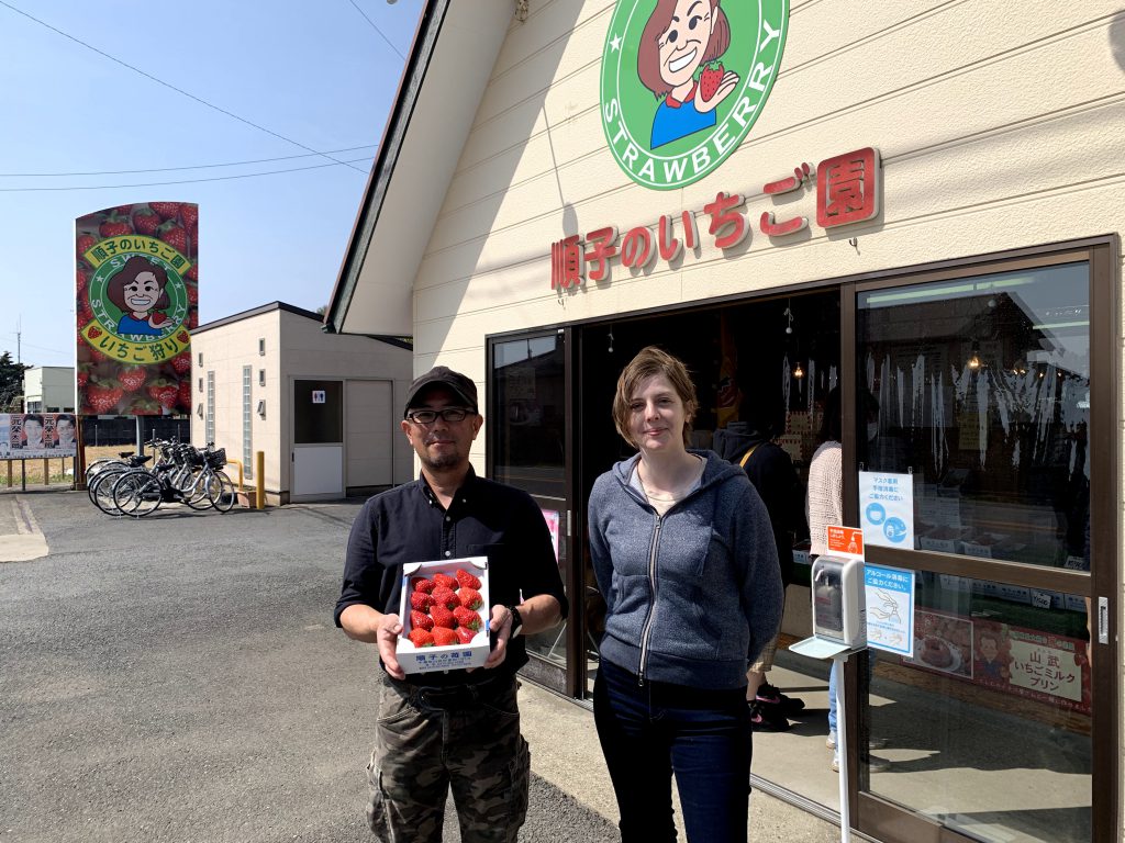 The author is standing next to Mr. Nakamura who is holding a pack of strawberries.