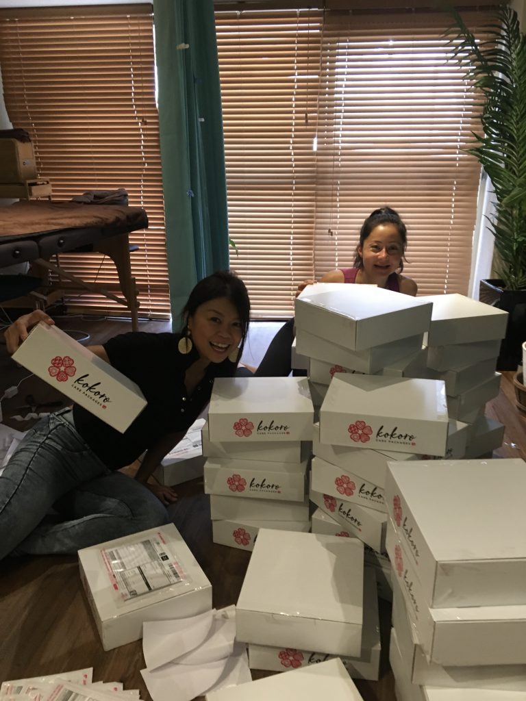 Two women surrounded by boxes with the 'Kokoro Care Package' logo