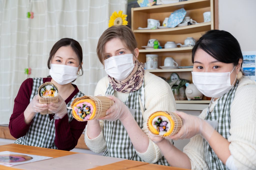 Three women use both hands to hold big sushi rolls