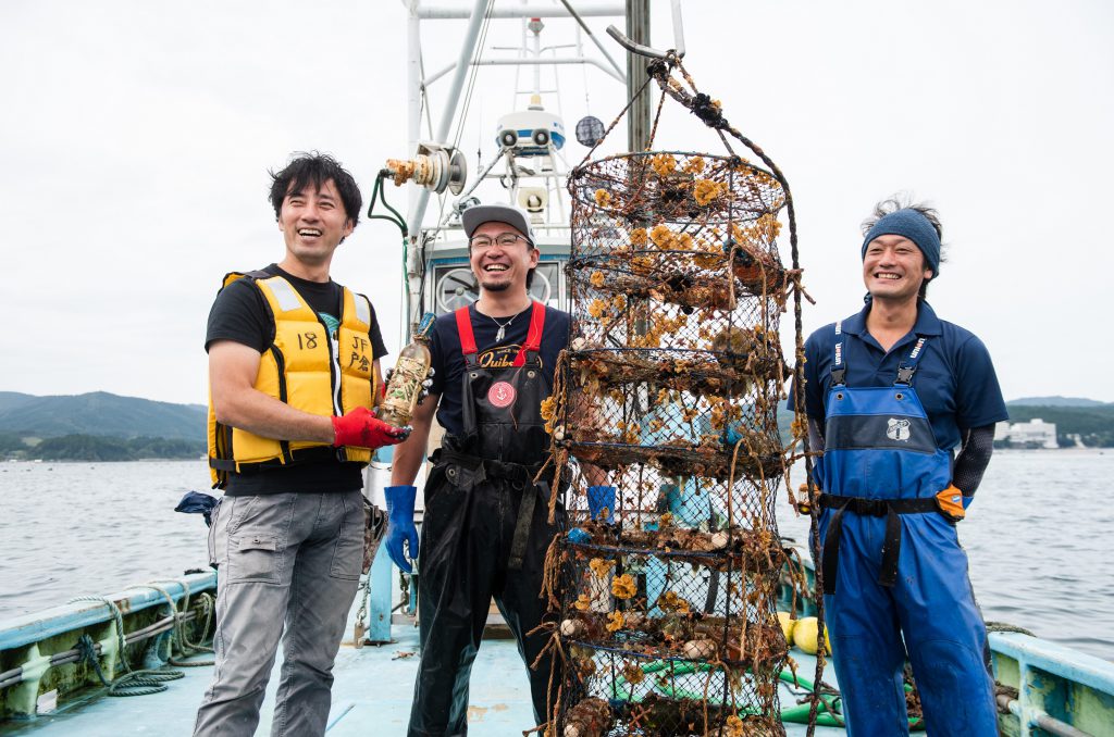 Mr. Sasaki is on a boat with two oyster producers. He is holding a bottle of wine. Next to them, a oyster net.