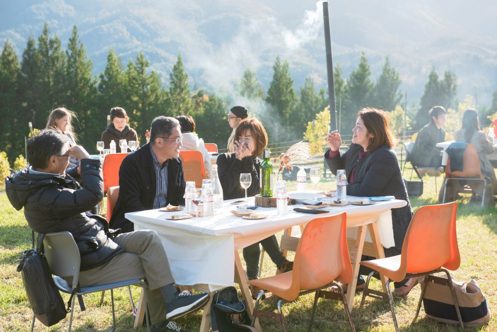 People at an open air gathering. They are sat around white clothed tables on which there are plates and glasses of wine.