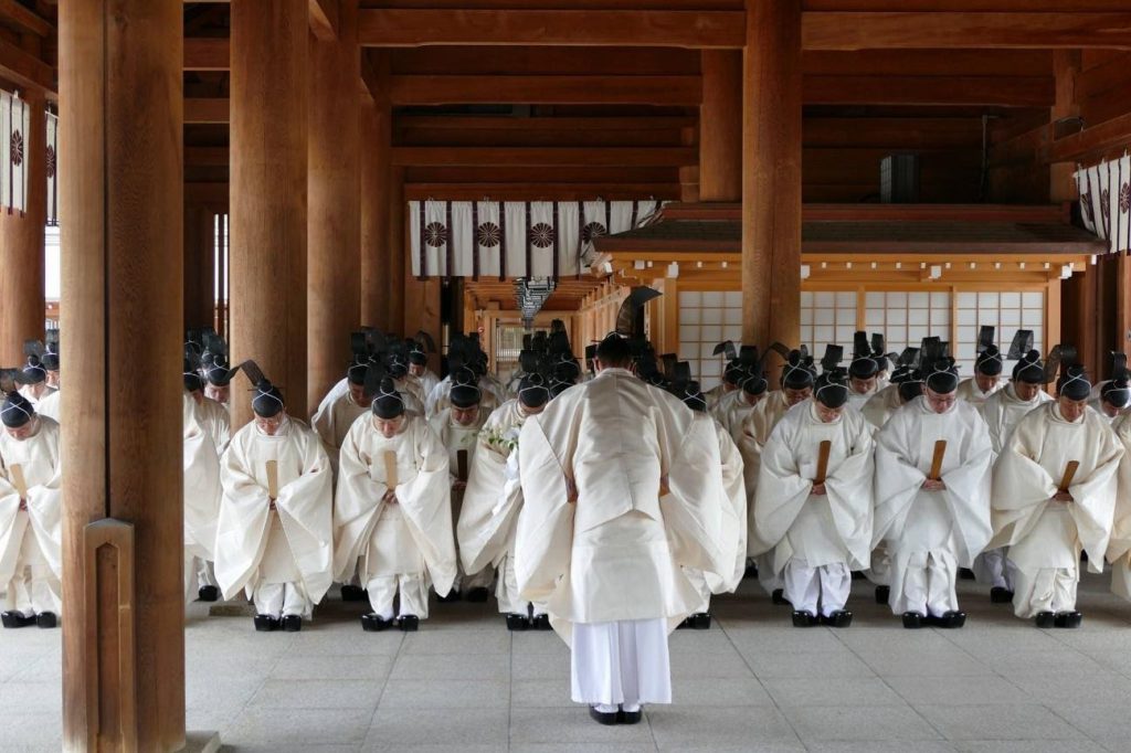 Inside a shrine, many Shinto priests dressed in white attire bow in front of a head priest.