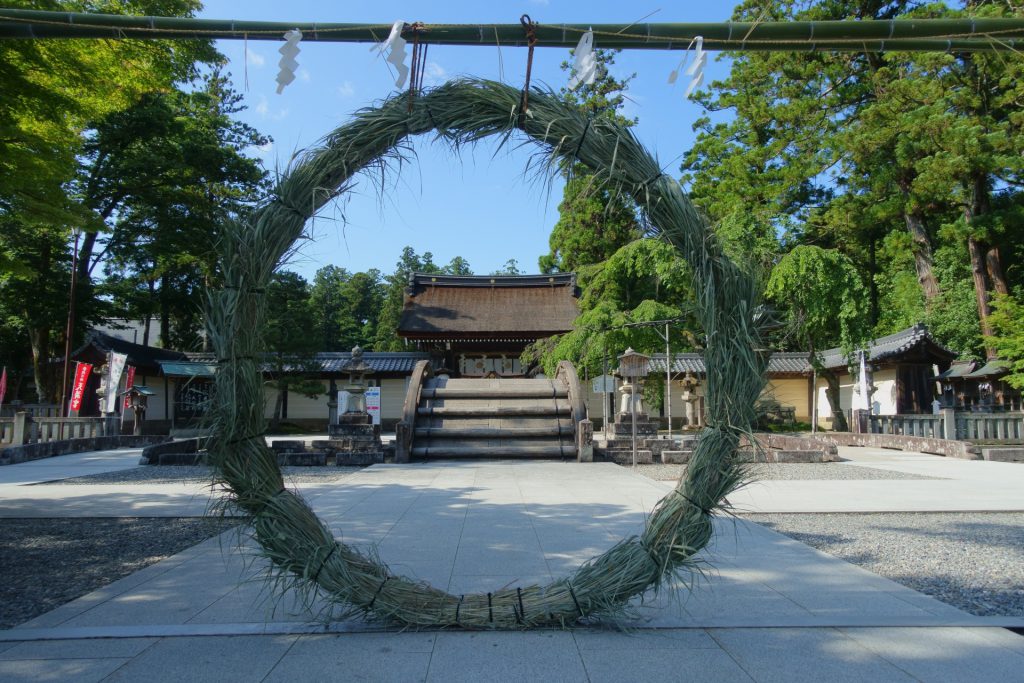 A huge straw ring large enough for a person to enter it is displayed vertically in front of a shrine.