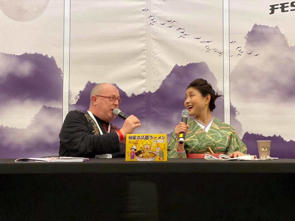 Cyril Coppini and Kikuhime Hayahsiya are sitting behind a desk and speaking in microphones.