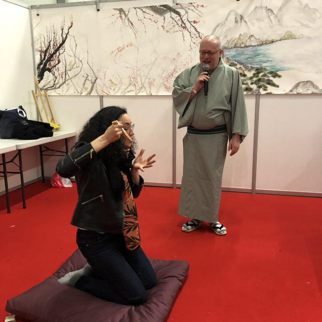 Cyril Coppini is standing next to a woman who is sitting on a cushion. She is miming eating food using a folding fan.