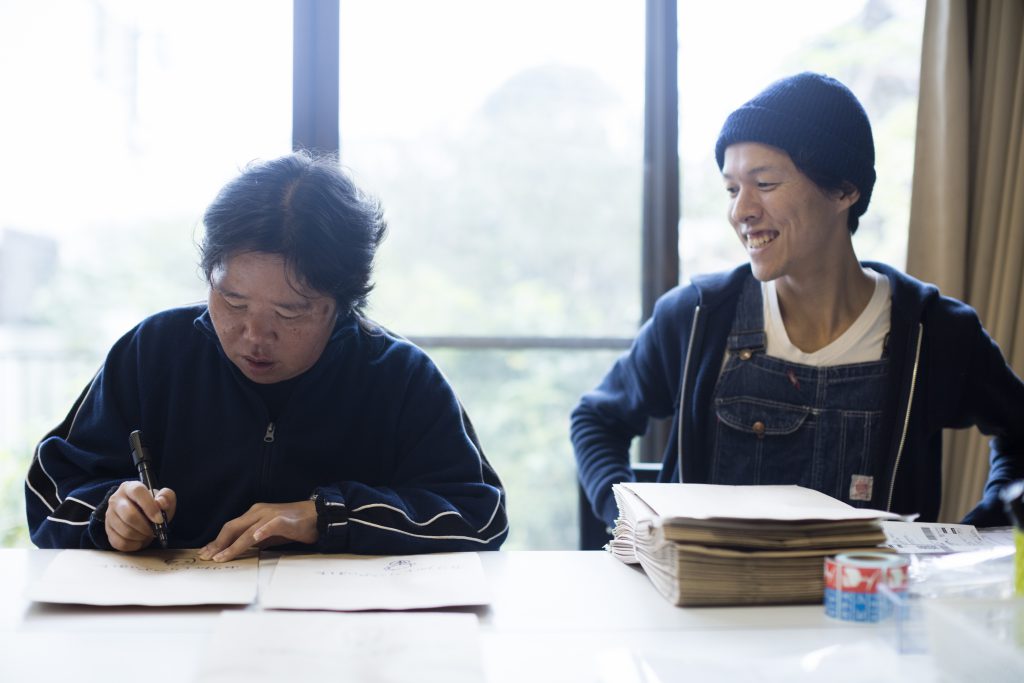 Go is smiling, sitting next to a woman who is writing on a paper