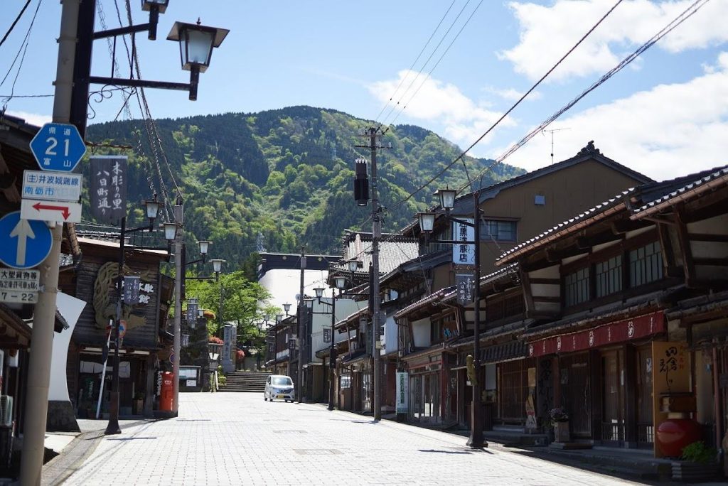 A view of a street in Inami. two-stories wooden houses are aligned on each side of the street. A mountain can see near the town.