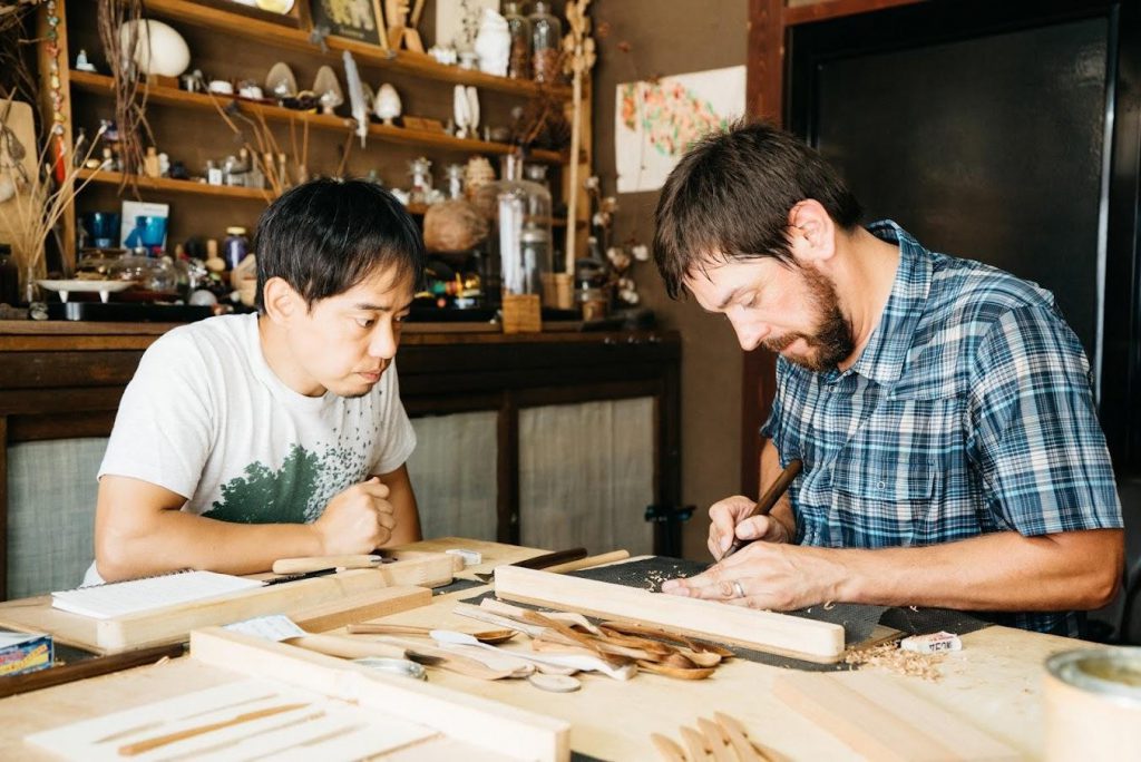 Komei Tanaka is looking at a Western guest carving wood.