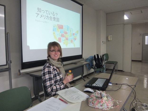 Kelsey Lechner, CIR with the JET program, is sitting at a desk in a classroom. Behind her, a screen shows a map of the United States.