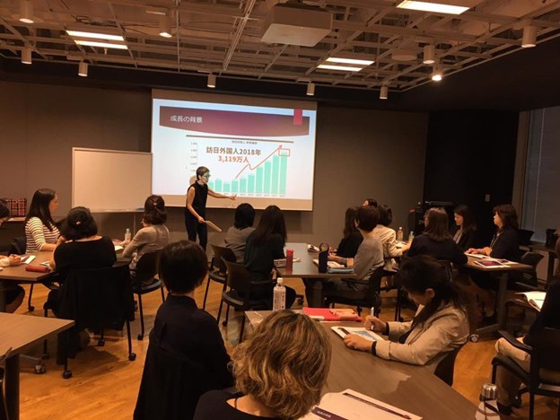 Cynthia Usui is standing in front of a screen, teaching. Women sitting in groups are listening to her.