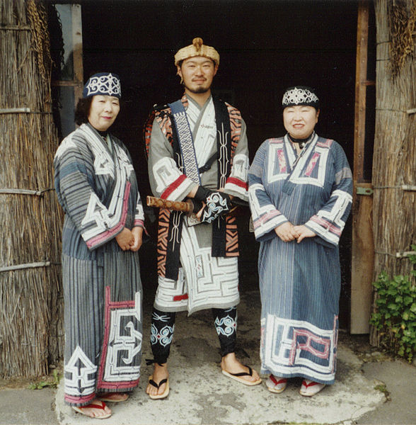 Ainu people dressed in traditional clothes are standing in front of a house.