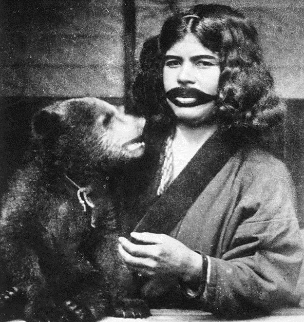 Black and white photo of an ainu woman. He mouth is tattooed and she is standing next to a baby bear.
