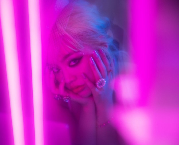 Chanmina's face is surrounded by pink neon lights. She looks straight at the camera with a serious look, both her hands holding her face.