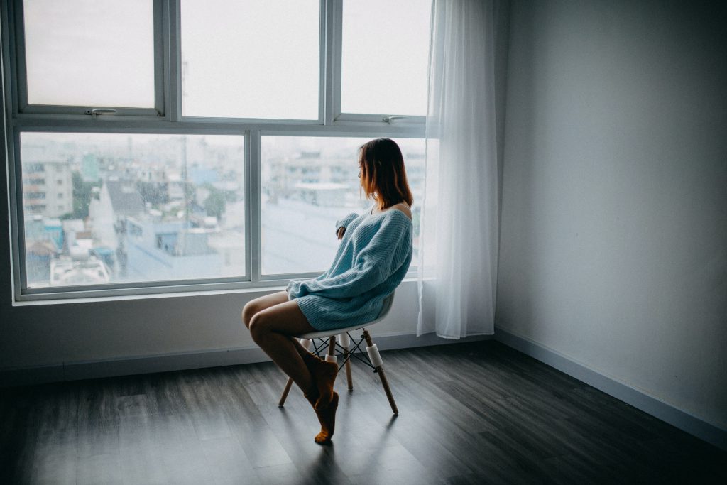 A woman is sitting alone on a chair by a window and looking down at the city.