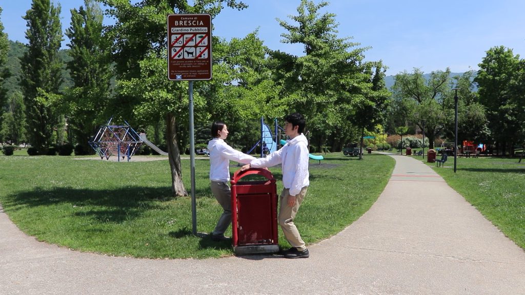 Aguyoshi, dressed in white jackets and beige trousers, are standing on each side of a trash bin in the middle of a park. A sign next to them shows they are in Bescia.