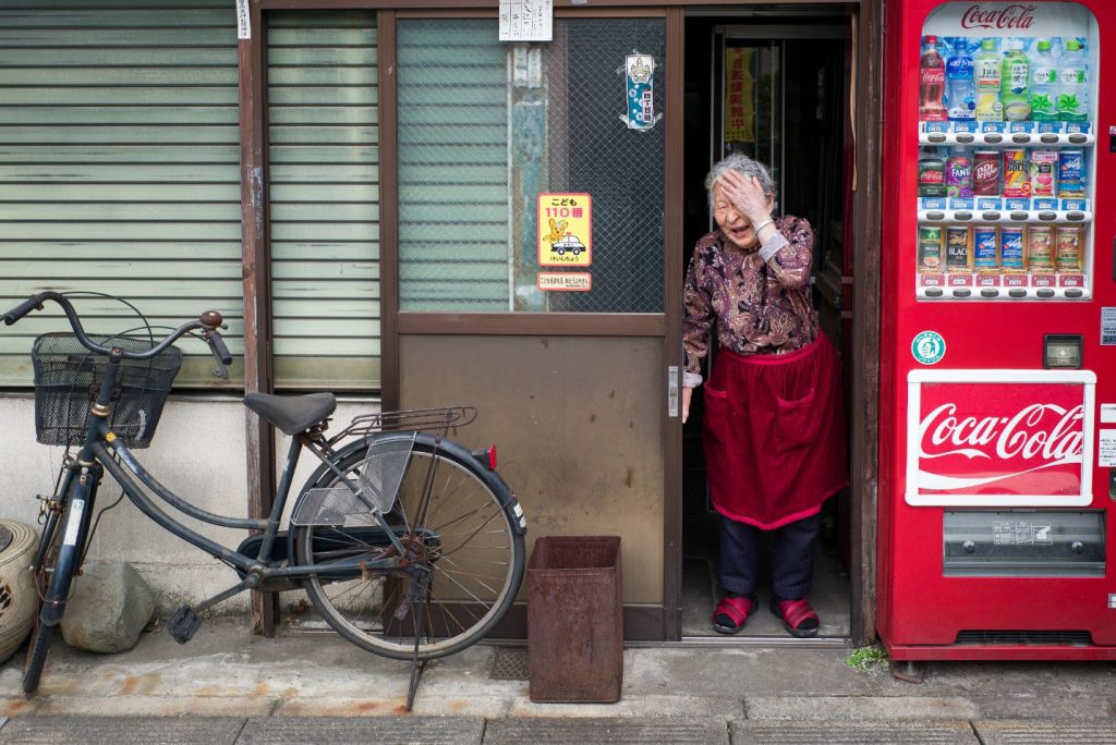 A picture taken by Lee Chapman shows an elderly woman at her front door. She is laughing and hiding half of her face with her hand. A red vending machine is next to her right. 