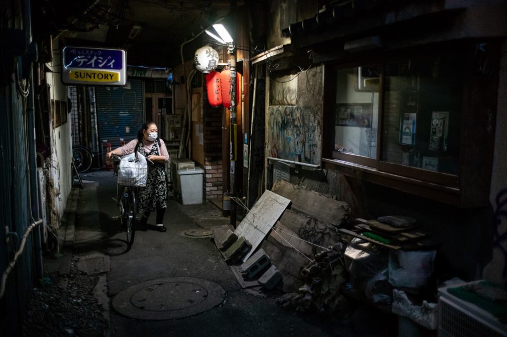 A picture taken by Lee Chapman shows a masked Japanese woman pushing her bicycle in a backstreet of Tokyo. The street is dimly lit and there are several discarded planks and garbage bins.