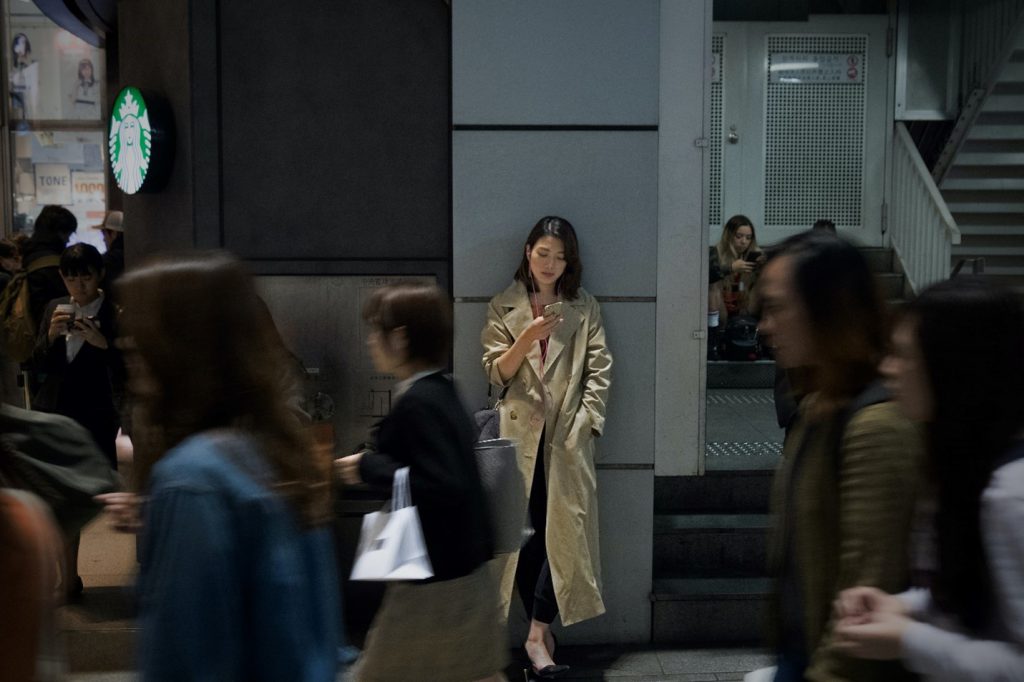 A woman is looking at her smartphone, her back on the wall in a busy Japanese street