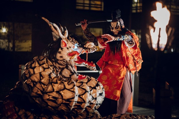 A Japanese traditional performance shows an actor dressed in orange traditional garment menacing a dragon with a sword.