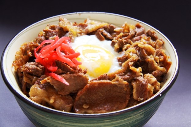 A bowl of rice on top of which are placed seasoned beef slices, marinated ginger, and an egg in the middle.