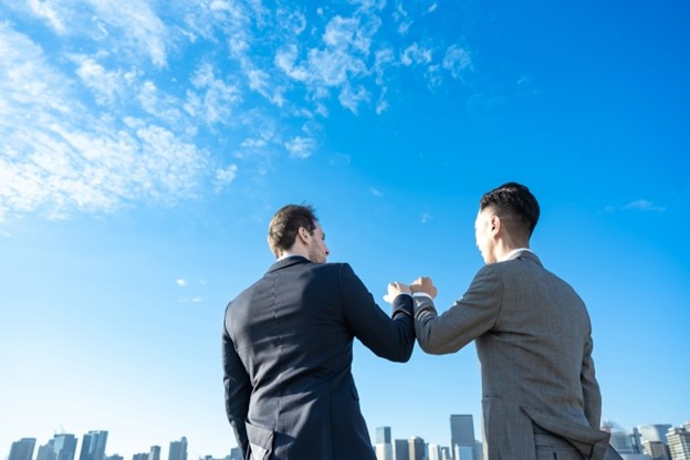 A Caucasian businessman and a Japanese businessman fist-bumping. Above their heads, the sky is blue.