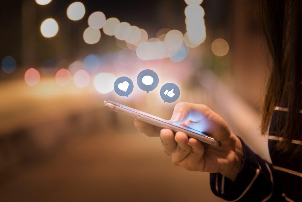 A person is holding a smartphone. On top of their hand that seems to be browsing float icons representing a heart, a speech bubble, and a thumbs-up.