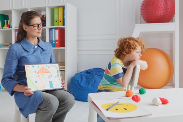 A woman holding a colorful drawing is shunned by a little boy who is sitting next to her but looking elsewhere.