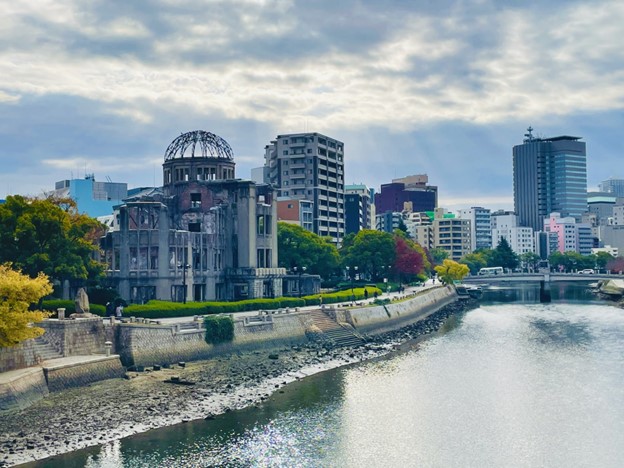 the picture is taken from a bridge above a river. The right half of the picture shows the river, with another bridge further away, and some tall concrete buildings behind it.
On the left half of the picture, there is a park with green trees and bushes, in the middle of which there is a concrete structure with empty windows and an empty dome at the top. It is the Genbaku dome, a building that remained after the Hiroshima atomic bomb.