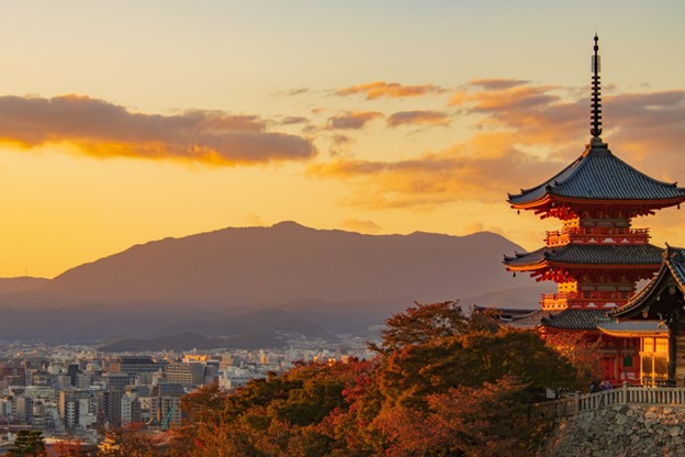 The picture is taken at sunset and the sky is a golden yellow. Far away, small mountains look purple. On the right of the picture, there is a three-story red pagoda surrounded by trees. It is at the top of a mountain, and overlooks the city below: we can see tall, concrete buildings.