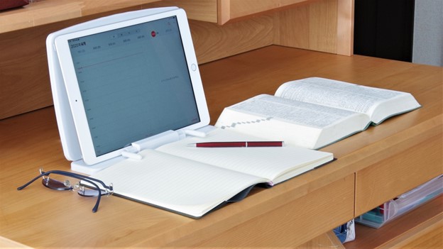 A tablet, a notebook, a paper dictionary, a pen and glasses lay on a wooden desk.