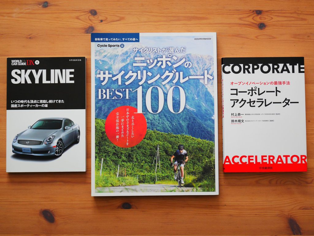 Three Japanese books lay on a wooden table. On the left, a world car guide called "Skyline" has a cover showing an elegant grey car. In the middle, the book with the highest format has a cover showing a cycler behind whom we can see trees and magnificent mountains. The title is "The best 100 cycling spots in Japan." The last book on the right has white cover with text only, in English and Japanese, that says "Corporate Accelerator."