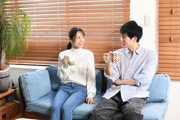 A young Japanese woman and a young Japanese man dressed in casual clothes are sitting on a blue sofa. They both hold coffee cups and seem to be chatting happily.