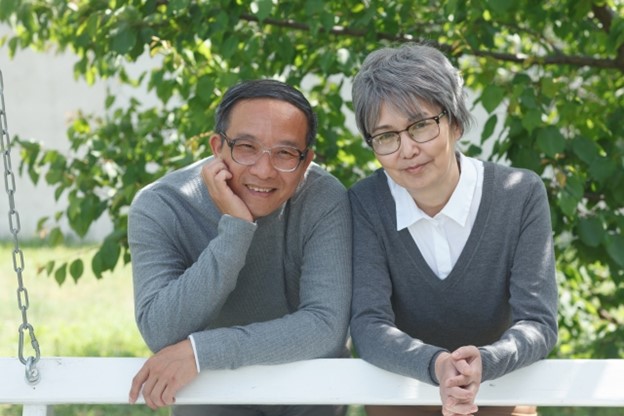 An elderly Japanese couple is smiling at the camera. They both wear glasses and a grey sweaters. They are in a garden, green tree branches behind them. They are leaning on a white barrier.