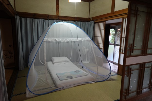 A traditional Japanese room with tatami floors and paper partitions. A mosquito net is set above a single person futon on the floor.