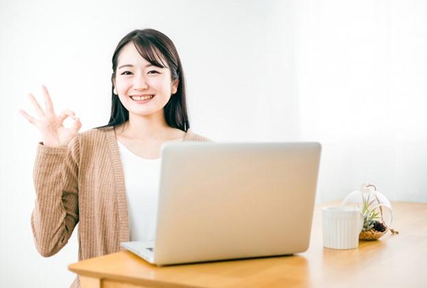 A Japanese young woman with long black hair is sitting behind her  grey laptop computer. She is wearing a brown cardigan. She is smiling at the viewer and is doing the ok sign with her right hand.