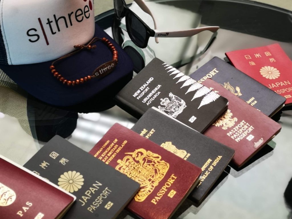 8 passports of different nationalities (Japan, New Zealand, India, United Kingdom) lay on a  glass table next to a cap and a pair of sunglasses.