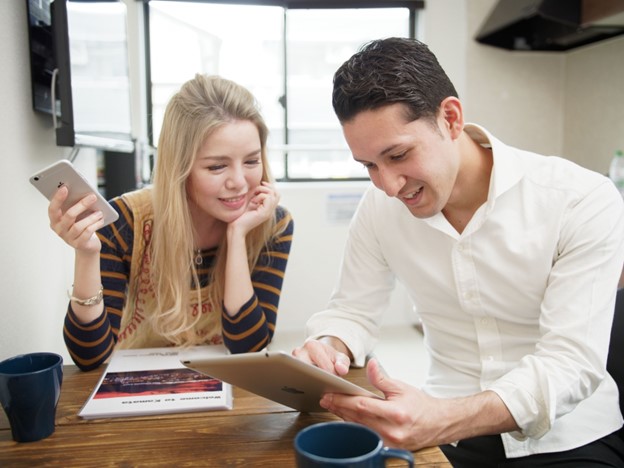 A blond Japanese woman and a caucasian man are sitting at a kitchen table, in front of cups of coffee. The woman is holding a smartphone and the man is using a tablet. They seem to be talking using these tools as a help for communication.