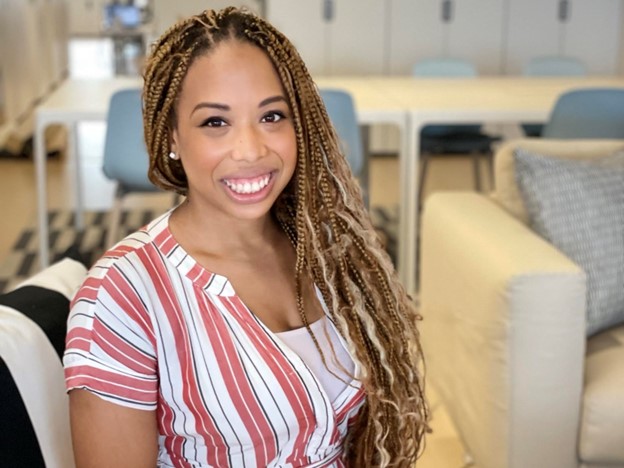 A picture Tiffany Rhodes smiling. Her hair is blonde and braided and she is wearing  a pink and white striped shirt. She is sitting in what seems to be a cosy open space office.