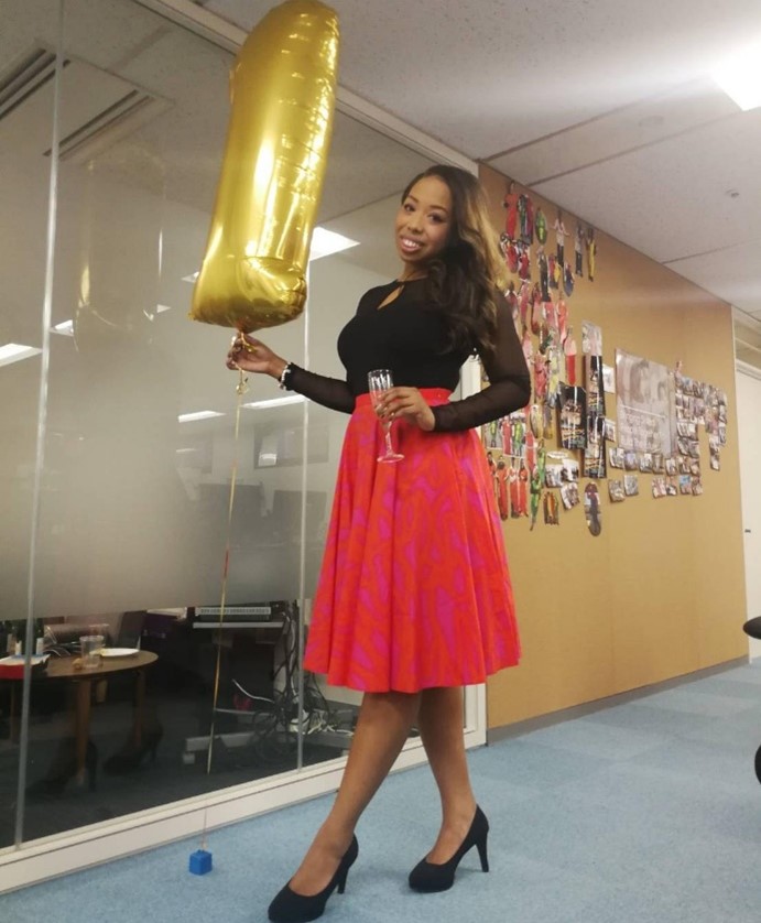 A body picture of Tiffany, smiling. She is wearing an elegant black shirt and a red long skirt, and black high-heel shoes. In her right hand, she is holding a golden balloon. In her left hand she has a glass of champagne.
The picture has been taken inside an office.
